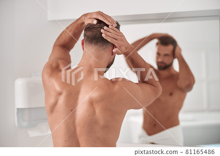 Sportive man strokes his healthy hair looking in large mirror in bathroom Sportive man strokes his healthy hair looking in large mirror in bathroom 81165486