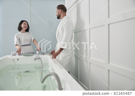 Woman in uniform shows hydro massage bath to man guest in modern salon Woman in uniform shows hydro massage bath to man guest in modern salon 81165487