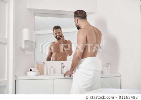 Seductive man with beard applies cream onto face in contemporary light bathroom Seductive man with beard applies cream onto face in contemporary light bathroom 81165488