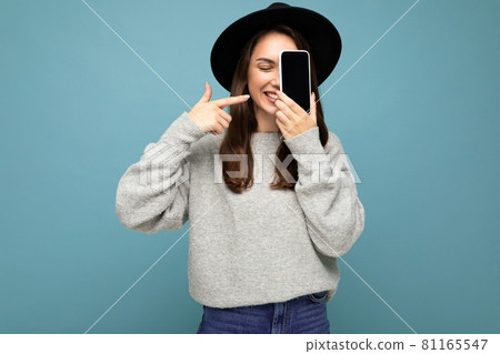 Photo of Beautiful positive woman wearing black hat and grey sweater holding mobilephone showing Photo of Beautiful positive woman wearing black hat and grey sweater holding mobilephone showing 81165547