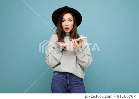 Beautiful young asking dissatisfied brunette woman wearing black hat and grey sweater holding 81166767
