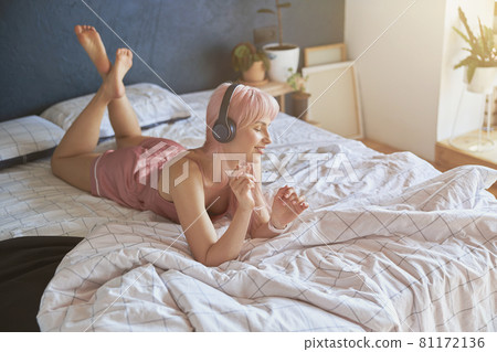 Stylish woman wearing pink silk pajamas enjoys with headphones lying on comfortable bed Stylish woman wearing pink silk pajamas enjoys with headphones lying on comfortable bed 81172136
