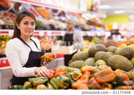 Female supermarket employee in apron lays fresh bell peppers on counter Female supermarket employee in apron lays fresh bell peppers on counter 81173406