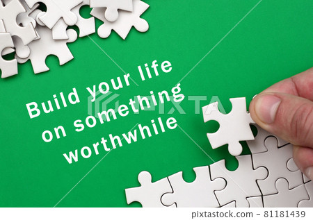Build your life on something worthwhile. The hand folds a white jigsaw puzzle and a pile of uncombed puzzle pieces lies against the background of the green surface 81181439