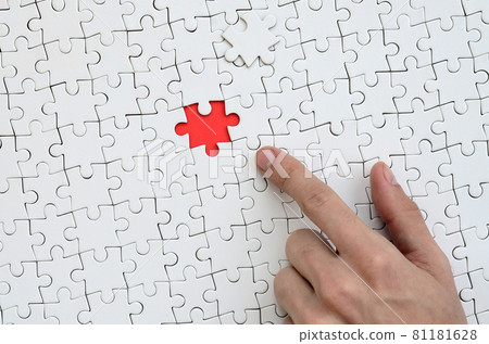 The texture of a white jigsaw puzzle in the assembled state with one missing element, forming a red space, pointed to by the finger of the male hand The texture of a white jigsaw puzzle in the assembled state with one missing element, forming a red space, pointed to by the finger of the male hand 81181628
