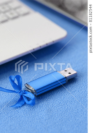 Brilliant blue usb flash memory card with a blue bow lies on a blanket of soft and furry light blue fleece fabric beside to a white laptop and smartphone. Classic female gift design for a memory card 81182544
