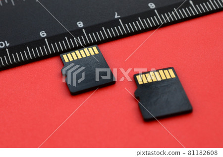 Two small micro SD memory cards lie on a red background next to a black ruler. A small and compact data and information store 81182608