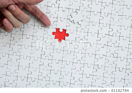 The texture of a white jigsaw puzzle in the assembled state with one missing element, forming a red space, pointed to by the finger of the male hand 81182784