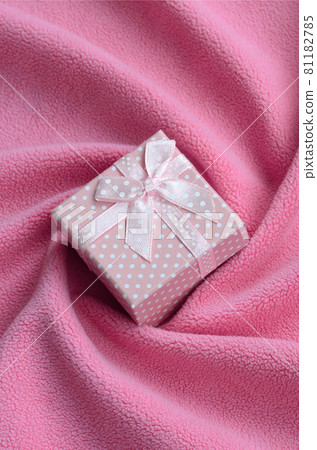 A small gift box in pink with a small bow lies on a blanket of soft and furry light pink fleece fabric with a lot of relief folds. Packing for a gift to your lovely girlfriend 81182785