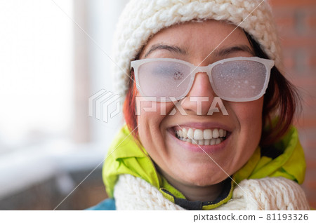 Smiling Caucasian woman stands on the balcony of a brick house in winter. Happy girl in ice-covered glasses on the street outdoors 81193326