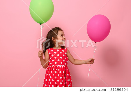Beautiful gorgeous 4 year pretty birthday girl in pink polka dot dress, holds two multicolored balloons, rejoices looking at a green balloon in her hand, isolated on pink background with copy space. 81196304