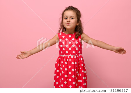 Smiling happy little charming girl with outstretched hands in a pink dress with polka dots, looking at the camera, posing on a pink background. Portrait of good-natured joyful, affable, friendly child 81196317