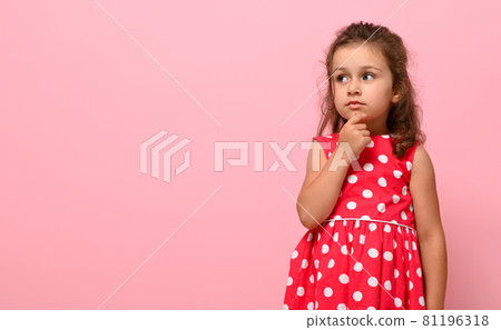 A cute adorable girl of preschool age, 3-4 years old, looks thoughtfully to the side , holding her finger at her chin, posing on a pink background with copy space 81196318