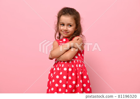 Gorgeous shyly 4 years kid in polka-dots dress hugs herself, sweet smiles posing on pink wall background with space for text. Confident portrait of beautiful baby girl for advertising Gorgeous shyly 4 years kid in polka-dots dress hugs herself, sweet smiles posing on pink wall background with space for text. Confident portrait of beautiful baby girl for advertising 81196320