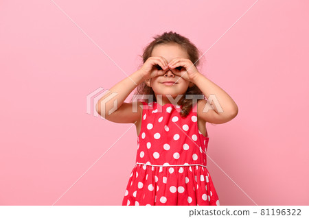 Smiling preschool girl in a pink polka dot dress, imitating a binocular with her hands and looking through it. Adorable child expressing positive emotions posing on pink background Smiling preschool girl in a pink polka dot dress, imitating a binocular with her hands and looking through it. Adorable child expressing positive emotions posing on pink background 81196322
