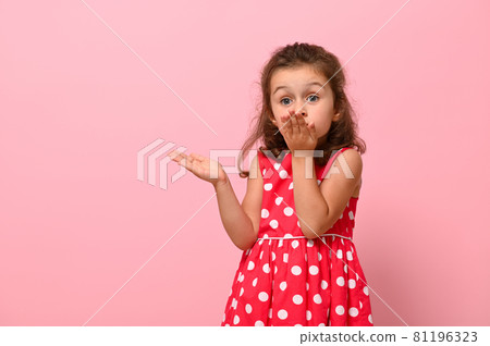 Gorgeous child girl in pink dress with polka dots sends an air kiss, looking at the camera, posing on a pink background with copy space. Portrait of a pretty kid with positive emotions for advertising Gorgeous child girl in pink dress with polka dots sends an air kiss, looking at the camera, posing on a pink background with copy space. Portrait of a pretty kid with positive emotions for advertising 81196323