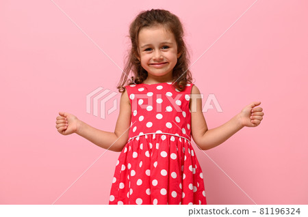 Face portrait of a beautiful child girl in pink polka dots dress, cute smiling looking at camera, posing with thumbs up, over pink background with copy space Face portrait of a beautiful child girl in pink polka dots dress, cute smiling looking at camera, posing with thumbs up, over pink background with copy space 81196324