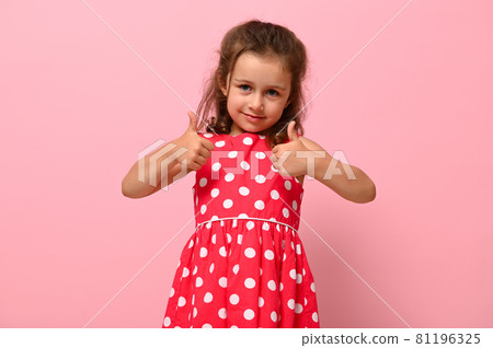 Portrait of beautiful child with positive emotions for advertising. Gorgeous baby girl in pink dress showing thumb up, cute smiling looking at camera, posing over pink background with copy space. 81196325
