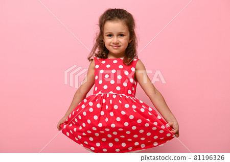 Portrait of cute charming girl in a pink dress with white polka dots posing onpink background cute smiling and holding the bottom of the dress with her hands. Beautiful child portrait for advertising Portrait of cute charming girl in a pink dress with white polka dots posing onpink background cute smiling and holding the bottom of the dress with her hands. Beautiful child portrait for advertising 81196326
