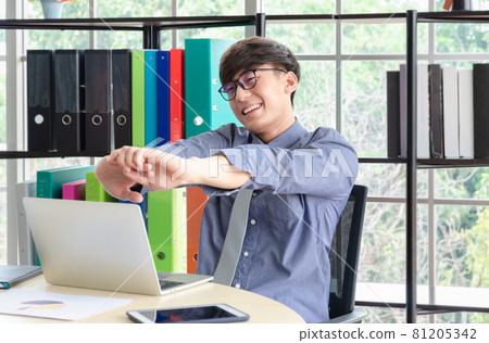 Cheerful young businessman sitting at office desk with laptop and stretching himself after finished work. Cheerful young businessman sitting at office desk with laptop and stretching himself after finished work. 81205342