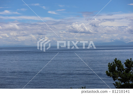 A landscape with a cloudy blue sky, green trees, a deep blue sea, and a vaguely hazy mountain on the opposite bank 81210141