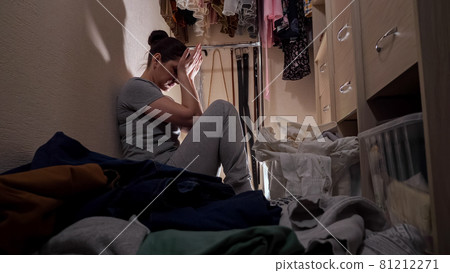 Woman suffering from hard depression sits near clothes pile Woman suffering from hard depression sits near clothes pile 81212271