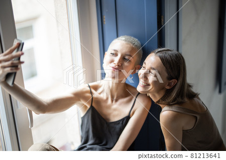 Two females having fun, talking online on cell phone by the window Two females having fun, talking online on cell phone by the window 81213641