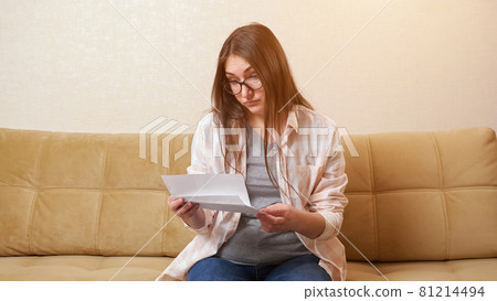 Woman opens envelope with bill and becomes nervous on couch 81214494