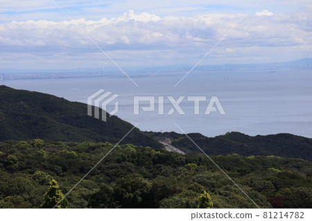A landscape with a cloudy sky, green mountains, roads, the sea, a vaguely hazy city on the opposite bank, mountains, and an artificial island with a bridge. 81214782