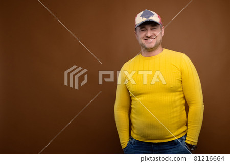 Portrait of an attractive friendly Caucasian man in a cap and yellow sweater on a brown background. Nice middle aged guy in casual clothes. Looks into the camera. Copy space 81216664