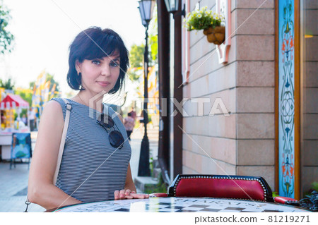 A brunette woman stands near a table of a street cafe in a European city on a busy street A brunette woman stands near a table of a street cafe in a European city on a busy street 81219271
