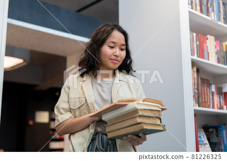 Happy girl with stack of books reading one of them Happy girl with stack of books reading one of them 81226325