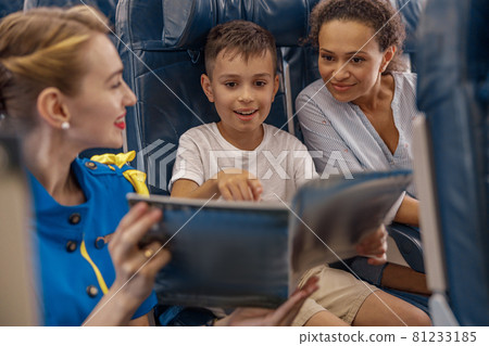 Female flight attendant entertaining a kid on board by offering a book to read. Cabin crew provide service to family in airplane Female flight attendant entertaining a kid on board by offering a book to read. Cabin crew provide service to family in airplane 81233185