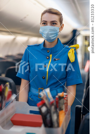 Stewardess in protective face mask looking away, serving food to passengers on aircraft. Air hostess walking with trolley on aisle 81233310