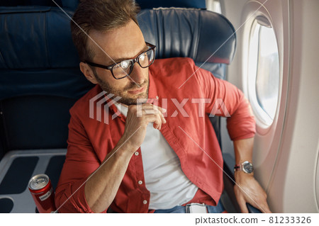 Handsome caucasian man in casual wear and glasses looking thoughtful, sitting on the plane near the window Handsome caucasian man in casual wear and glasses looking thoughtful, sitting on the plane near the window 81233326