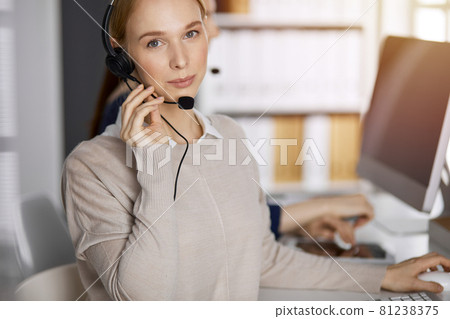 Friendly caucasian businesswoman talking by headset in sunny office. Call center and diverse people group in business Friendly caucasian businesswoman talking by headset in sunny office. Call center and diverse people group in business 81238375