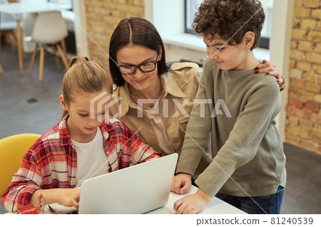 Experiment. Caring young female teacher in glasses smiling while helping smart kids to learn programming using laptop during STEM class Experiment. Caring young female teacher in glasses smiling while helping smart kids to learn programming using laptop during STEM class 81240539