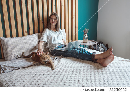 Young woman using laptop to work, lying on the bed with a pet cat in the bedroom, . Flexible hours and remote work. 81250032