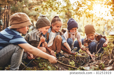 Happy school children boys and girls with backpacks making bonfire with magnifying glass in forest 81251919