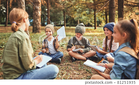 School children with notebooks and female teacher sitting on ground in forest during ecology lesson 81251935