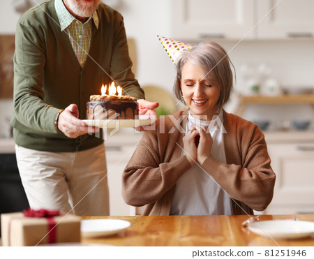 Happy elderly woman in party hat closing her eyes, she makes a wish, while her husband holds a chocolate cake with candles during Birthday celebration Happy elderly woman in party hat closing her eyes, she makes a wish, while her husband holds a chocolate cake with candles during Birthday celebration 81251946