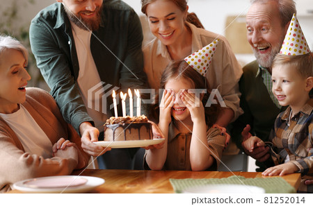 Happy big family surprising excited little girl by bringing Birthday chocolate cake with lit candles 81252014
