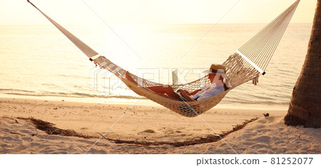 Young woman female freelancer using laptop while lying in hammock on the tropical beach at sunset Young woman female freelancer using laptop while lying in hammock on the tropical beach at sunset 81252077