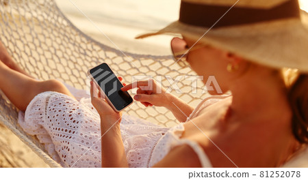 Working at sunset. Young happy female using smartphone while lying in the hammock on the beach Working at sunset. Young happy female using smartphone while lying in the hammock on the beach 81252078