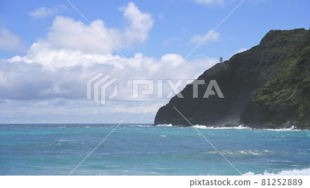 View of makapuu lighthouse. Waves of Pacific Ocean wash over yellow sand of tropical beach. Magnificent mountains of Hawaiian island of Oahu against backdrop of blue sky with white clouds. 81252889