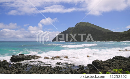 View of makapuu lighthouse. Waves of Pacific Ocean wash over yellow sand of tropical beach. Magnificent mountains of Hawaiian island of Oahu against backdrop of blue sky with white clouds. 81252892