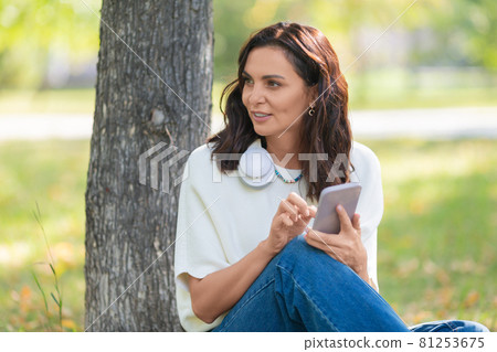 Photo of a 40-year-old woman with a smartphone sitting on a grass in city park. Photo of a 40-year-old woman with a smartphone sitting on a grass in city park. 81253675