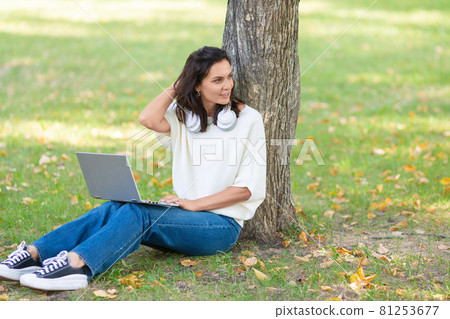 Photo of a 40-year-old woman with laptop sitting on a grass in city park. Photo of a 40-year-old woman with laptop sitting on a grass in city park. 81253677