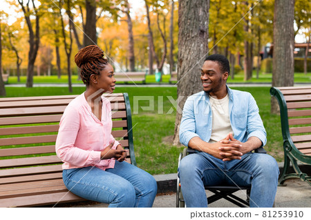 Cheerful disabled black guy in wheelchair having conversation with his girlfriend at park in autumn Cheerful disabled black guy in wheelchair having conversation with his girlfriend at park in autumn 81253910