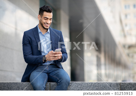 Successful IT manager sitting next to office building, using smartphone Successful IT manager sitting next to office building, using smartphone 81253913